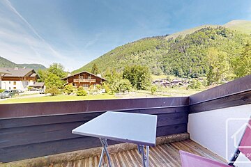 Balcony view of a green alpine landscape with a folding table, wooden houses, and lush mountains in the background under a blue sky.