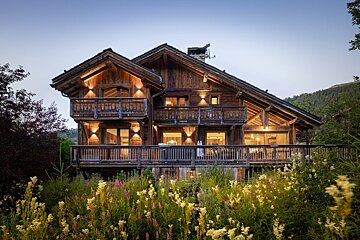 A large wooden house is surrounded by flowers and trees
