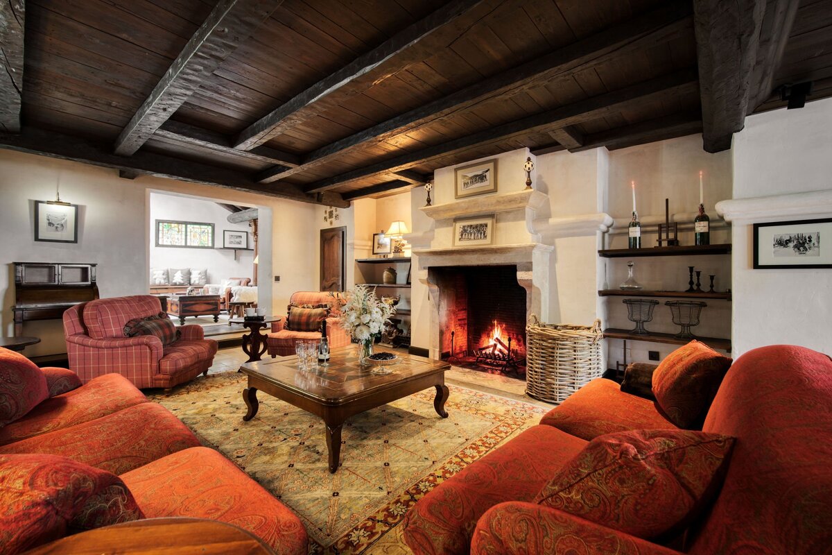 Warm, rustic living room featuring a cozy fireplace, dark wood-beamed ceilings, plush red sofas, and a central wooden coffee table.