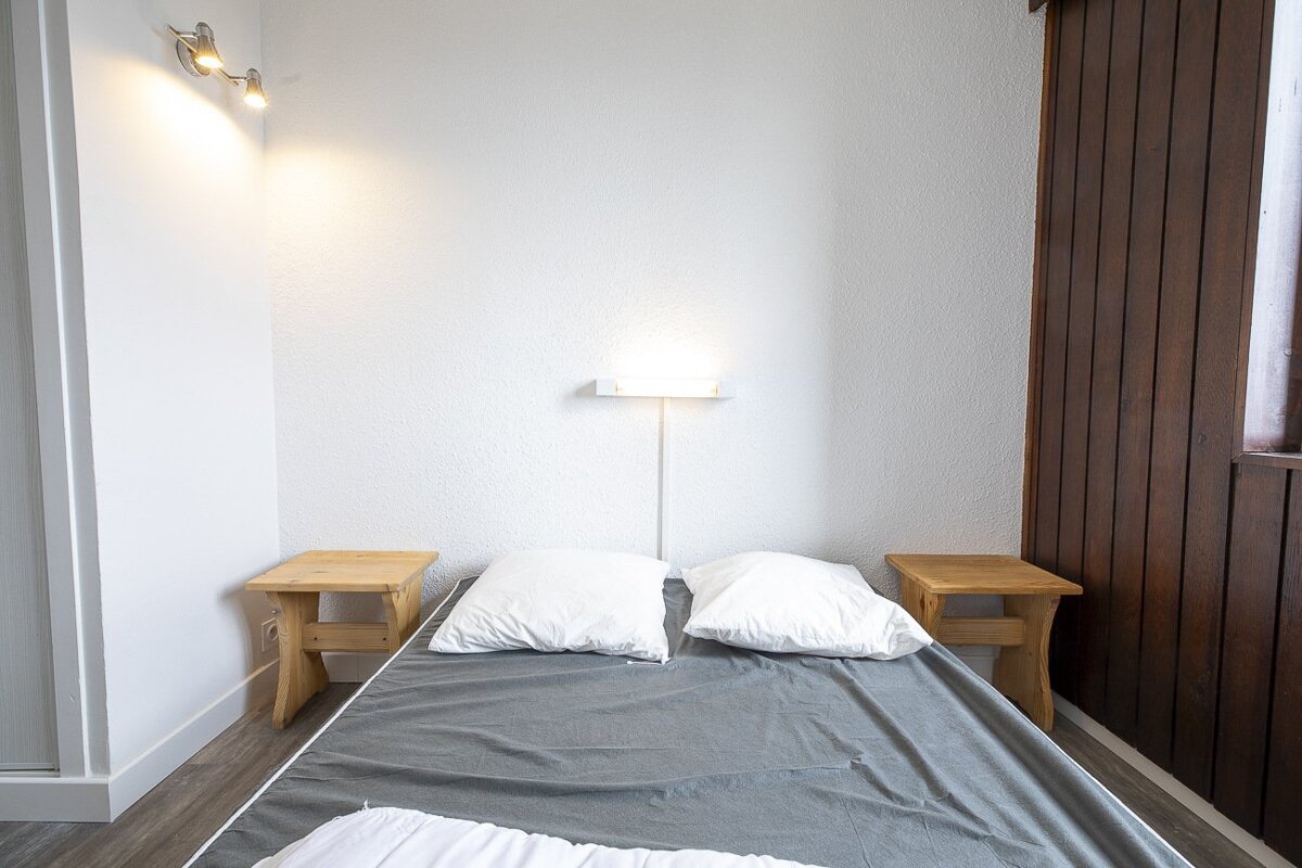 A minimalist bedroom with a bed, grey blanket, white pillows, and two small wooden stools. White walls, dark wood paneling, and bright lights.