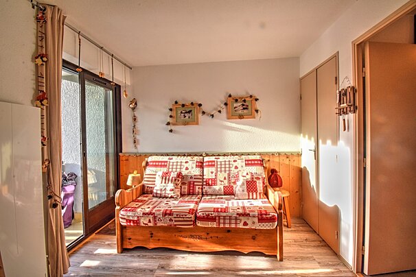 A cozy, sunlit room with a rustic wooden sofa featuring red/white patterned cushions. A balcony door is on the left, and simple decor adorns the walls.
