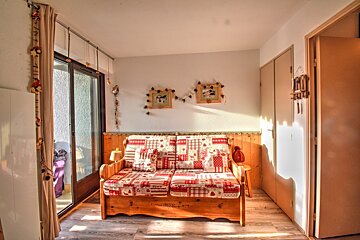 A sunlit, cozy room with a rustic wooden sofa, patterned cushions, and charming mountain-themed decor. A balcony door is visible on the left.
