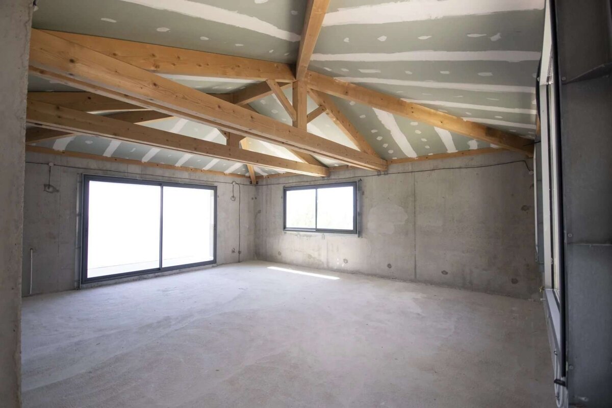 An unfinished room with concrete walls and floor, exposed wooden beams on a partially drywalled ceiling, and bright windows.