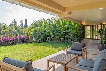 A covered patio with modern outdoor seating overlooks a vibrant green lawn bordered by tropical plants, purple flowers, and a fence under a bright sky.