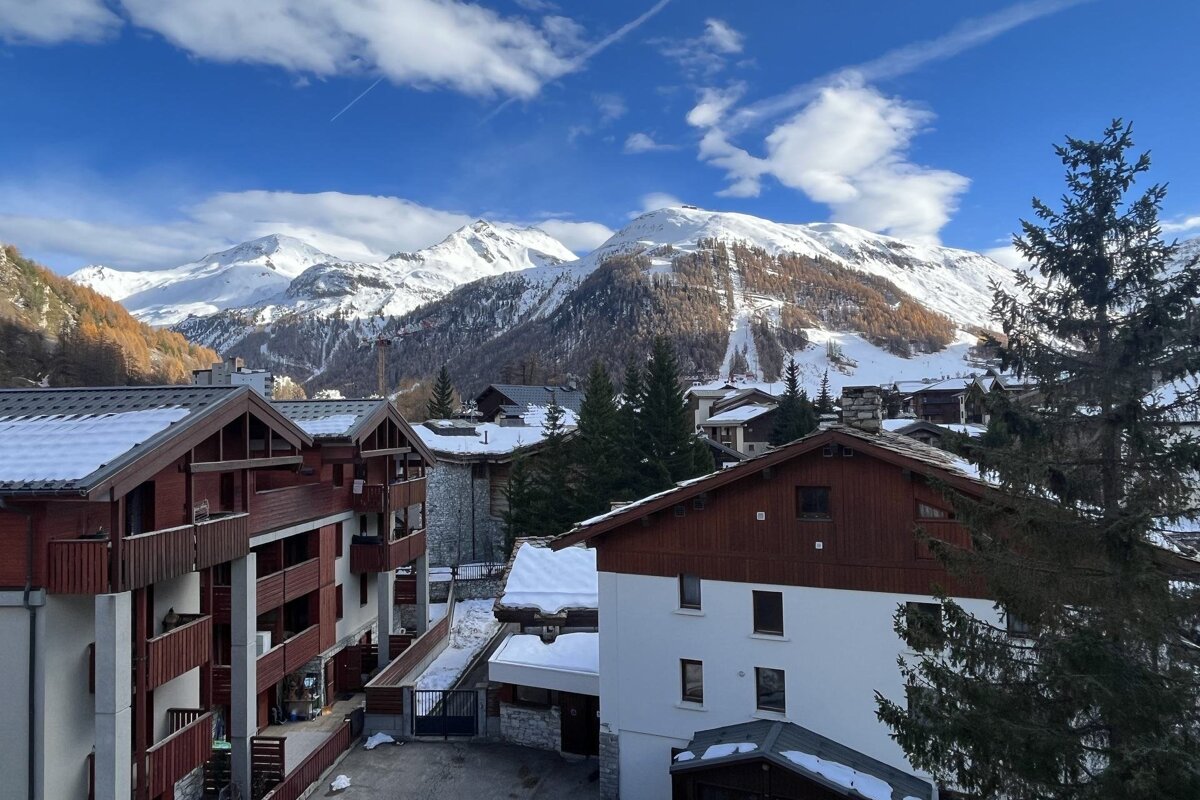 A sunny alpine village with snow-dusted buildings and pine trees, framed by vast snow-capped mountains under a bright blue sky.