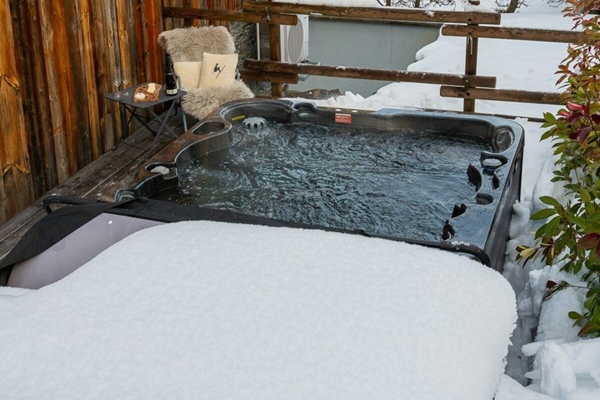 An outdoor hot tub steams on a snowy wooden deck next to a cozy chair with a fur throw and a small table with wine.