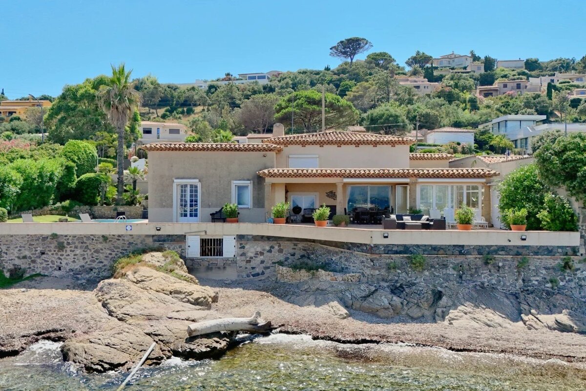 A sunny Mediterranean villa built on a rocky shoreline overlooking the sea, with a terrace and lush green hills in the background.