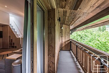 Wooden chalet balcony overlooking lush green trees, with an open door showing a modern interior living space.