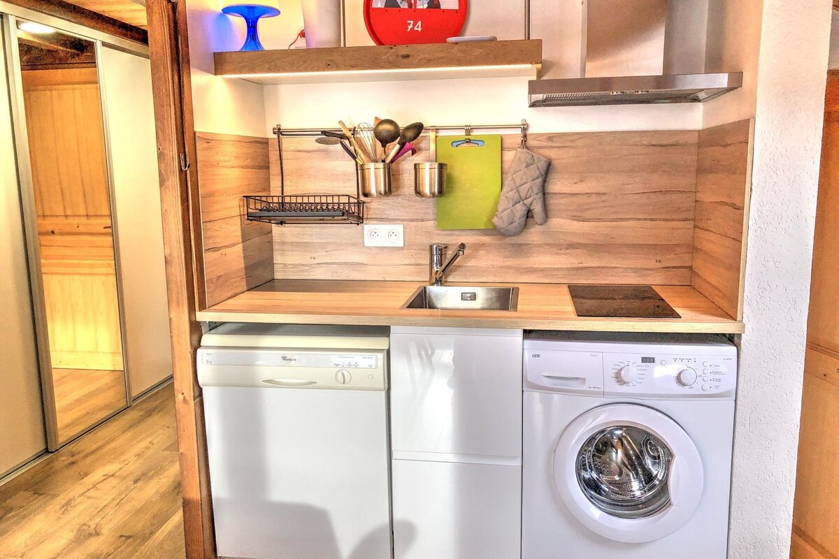 A compact kitchen with a sink, hob, dishwasher, washing machine, and wooden backsplash. Hanging utensils and a lighted shelf complete the functional space.