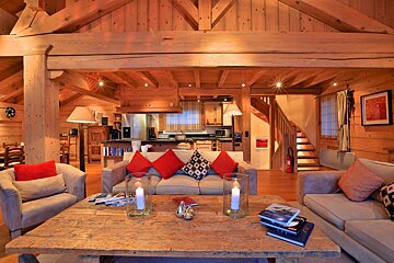 Warm, rustic wooden chalet interior with an open-plan living room, kitchen, and staircase. Features comfy sofas, a large coffee table with candles, and cozy lighting.