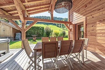 Inviting wooden patio with a dining table, hot tub, and exposed beams, offering stunning views of rolling green hills and distant forests.
