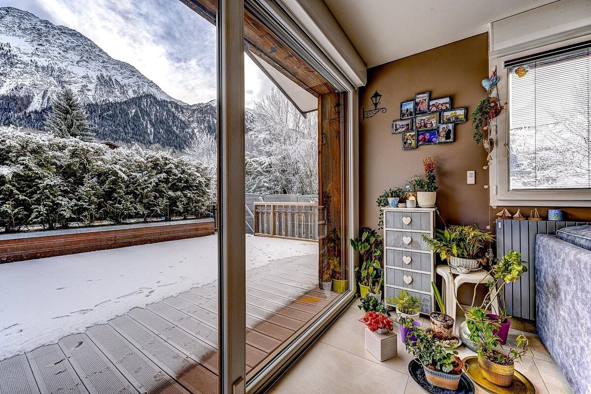 A cozy room with many plants and photos looks out through a large glass door onto a snowy patio and majestic, snow-covered mountains.