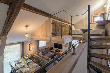 A multi-level rustic interior with a loft bedroom, spiral staircase, dining table, and living area. Features warm wood, exposed beams, and industrial metal accents.