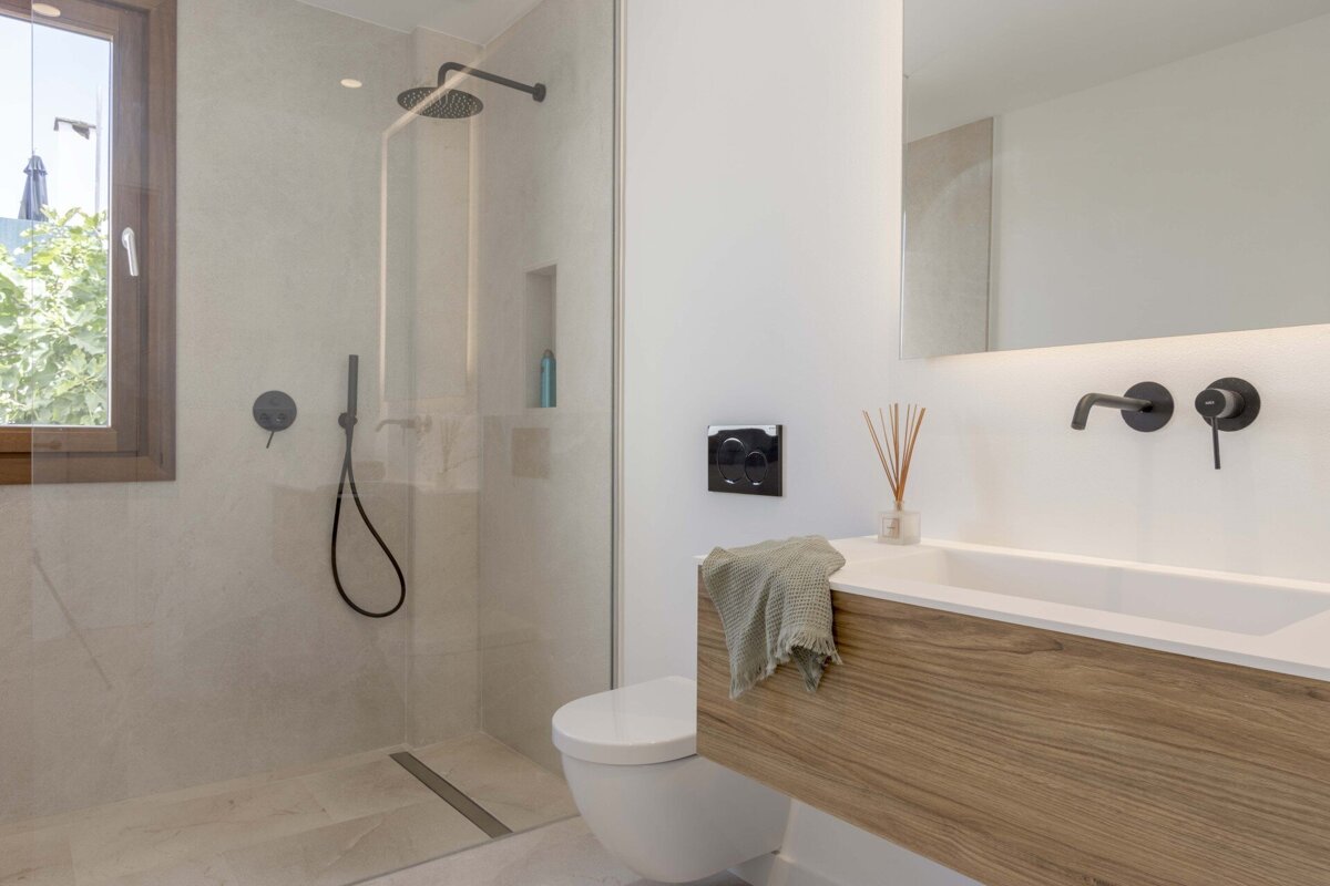 A modern bathroom featuring a glass-enclosed shower with black fixtures, a wall-mounted toilet, and a wooden vanity with a white sink and lit mirror.