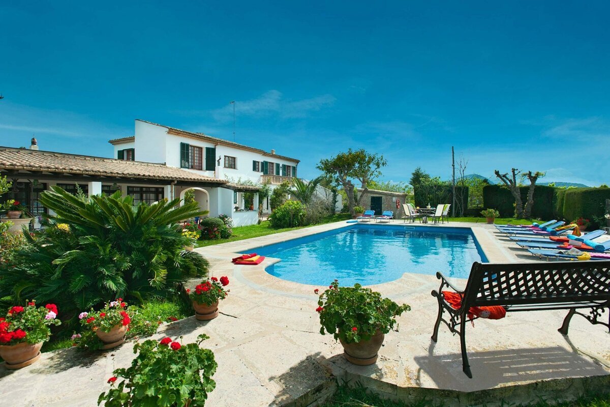A serene villa with a bright blue swimming pool, lush garden, potted flowers, and sun loungers under a clear blue sky.