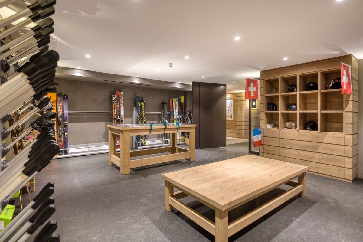 A well-lit, modern ski room featuring rows of skis and boots, wooden tables, and a wooden cabinet with helmets and Swiss flags.
