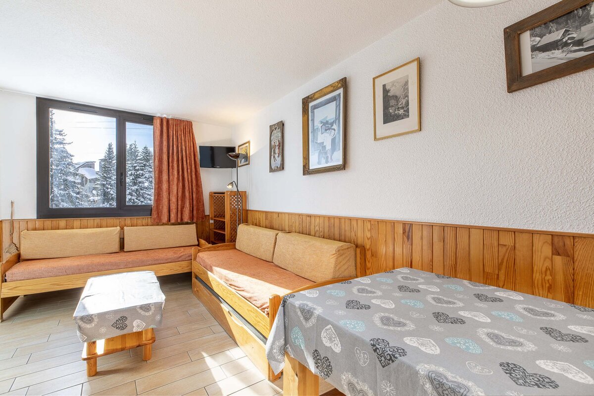 Cozy living room with wooden furniture, two sofas, and a dining table. A window offers views of a snowy landscape. Pictures adorn white walls.