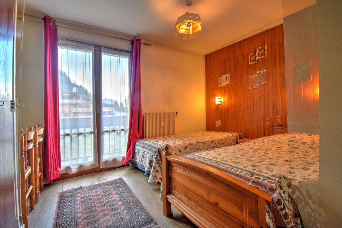 A warm bedroom features two wooden twin beds with patterned covers, a large window with red curtains, and wood paneling. A rug lies on the floor.