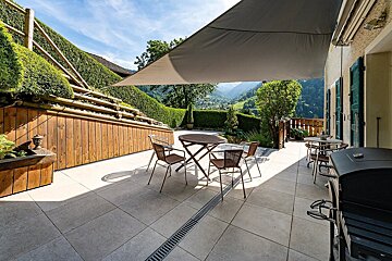 A sunny, shaded patio with tables, chairs, and a grill, framed by lush hedges and offering a picturesque view of a green mountain valley.