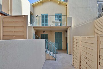 A cream-colored building with blue shutters, a balcony with furniture, and stairs. An outdoor patio is visible, framed by light-toned fences.