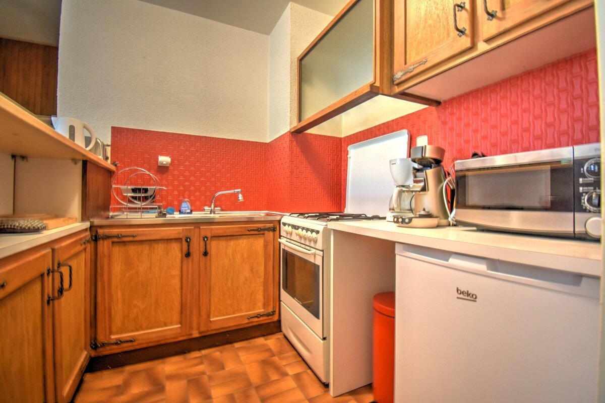 A functional kitchen with a vibrant red patterned backsplash, wooden cabinets, sink, stove, microwave, coffee maker, and a Beko fridge.