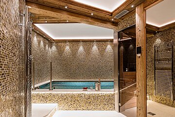 Luxurious indoor spa featuring a small pool, golden mosaic tiles, warm wooden beams, and an adjacent sauna room.