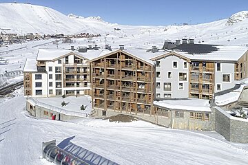 Modern alpine resort buildings with wood and gray facades nestled in vast snow-covered mountains under a clear blue sky, with ski slopes in the foreground.