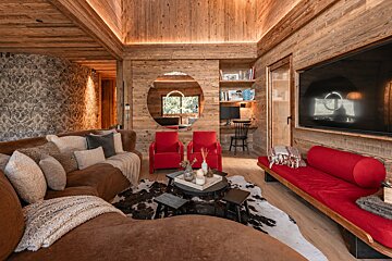 Cozy rustic living room with wood-paneled walls, featuring a brown curved sofa, red accents, a cowhide rug, and a large round mirror.