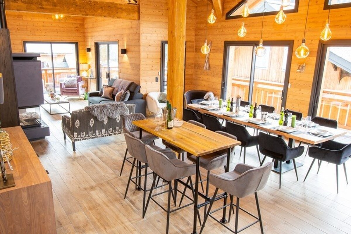 Cozy wood-paneled chalet interior with living and dining areas, warm hanging lights, and large glass doors.