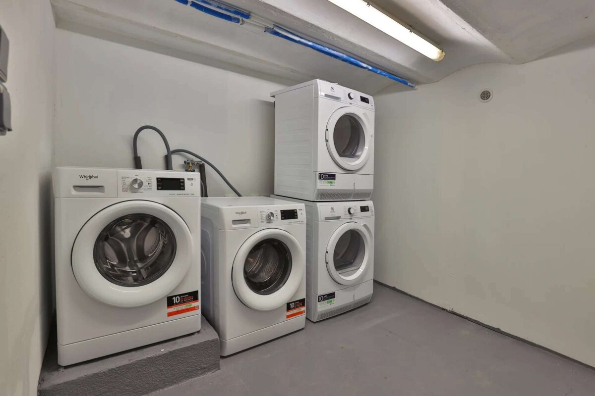 A clean, utilitarian laundry room featuring four white washing machines and dryers, some stacked, on a grey floor with white walls and blue pipes overhead.