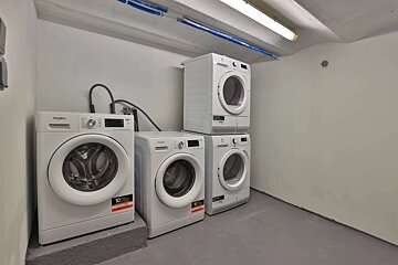 A clean, utilitarian laundry room featuring four white washing machines and dryers, some stacked, on a grey floor with white walls and blue pipes overhead.