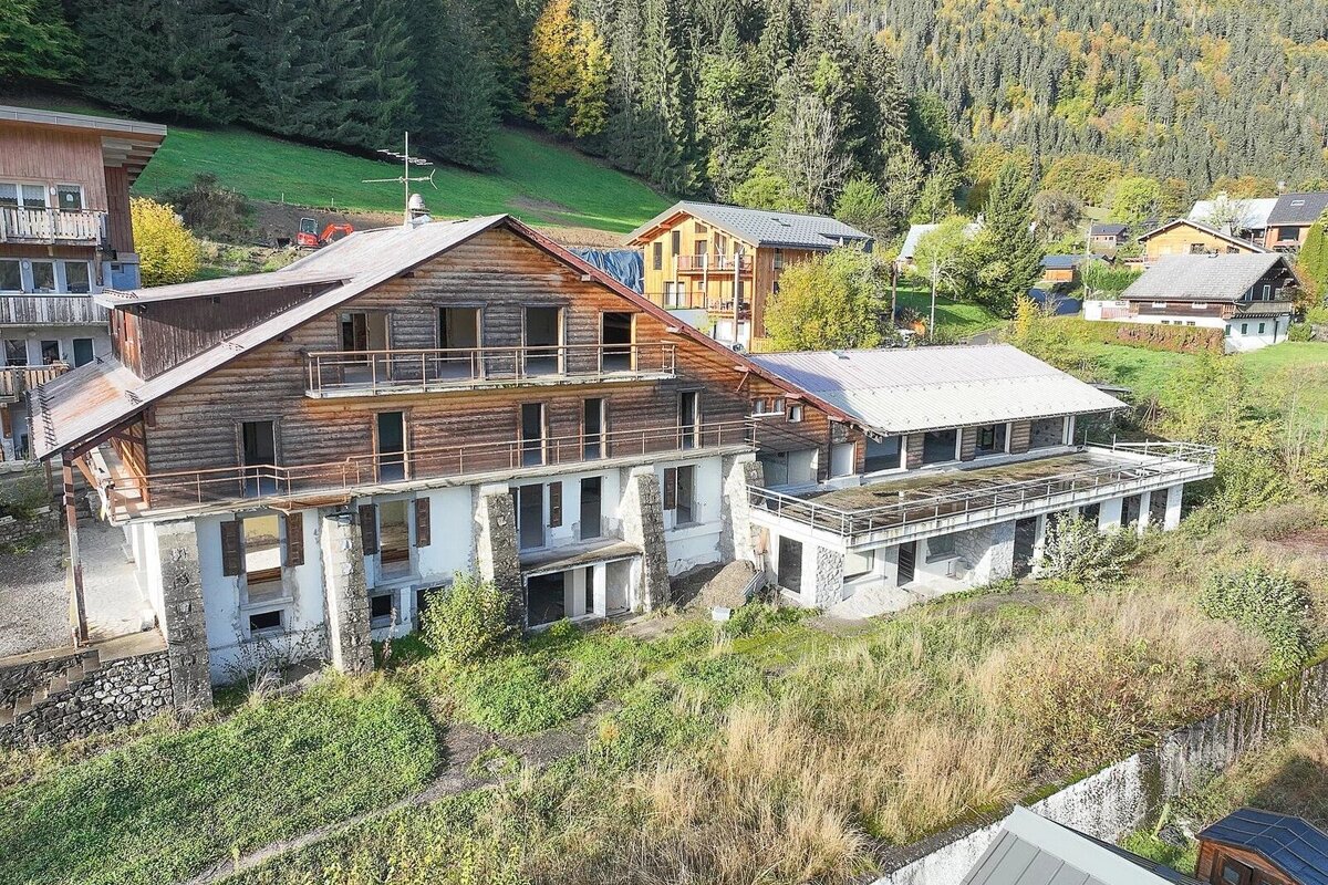 An aerial view of an old, multi-story wooden and stone building complex on a hillside, surrounded by overgrown grass and distant forest.