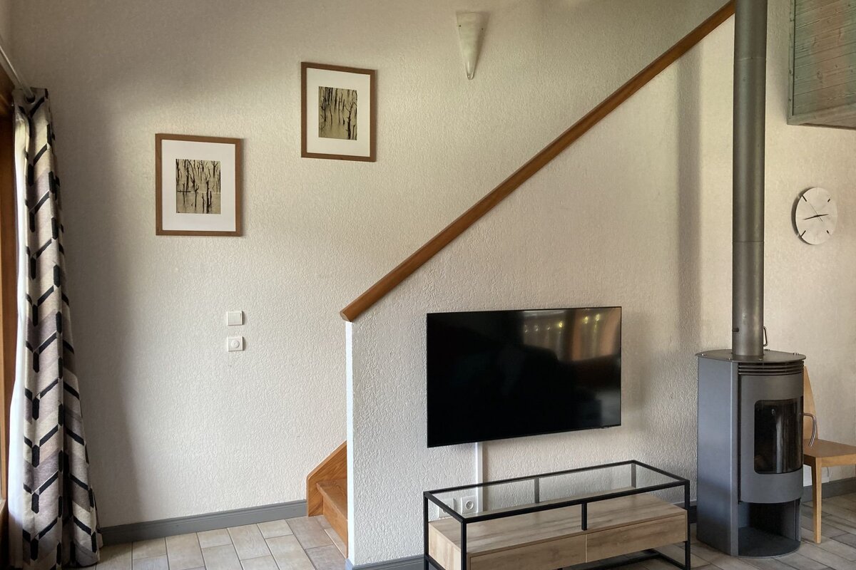 A living room with a wall-mounted TV, a glass media console, a modern wood-burning stove, and stairs leading up behind it.