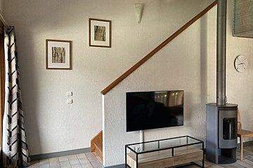 A living room with a wall-mounted TV, a glass media console, a modern wood-burning stove, and stairs leading up behind it.