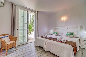 A bright, white bedroom features two single beds, a wicker chair, and a sliding door opening to a green garden. The room has light wood flooring.