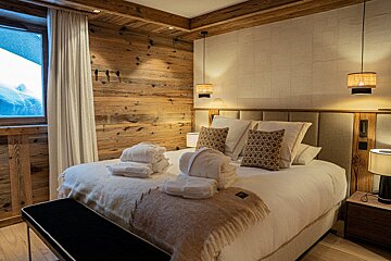 Cozy, rustic bedroom with wooden walls, a large bed with white linens and towels, and a window showing snow. Warm lighting creates an inviting chalet feel.