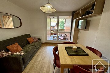 Cozy living/dining room with a dark green sofa, wooden dining table, TV, and a glass door leading to a balcony. Warm, modern decor.