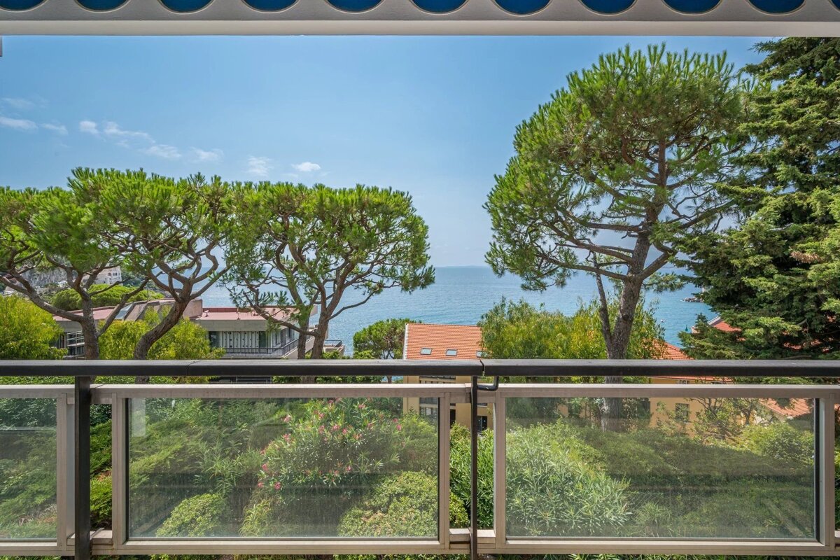 Balcony view of a serene blue sea, framed by several pine trees and lush green foliage, with distant houses.