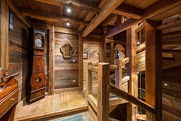 A cozy, rustic wooden interior with exposed beams, a grandfather clock, an ornate mirror, and a multi-level wooden staircase bathed in warm light.