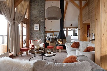 A cozy, modern chalet living room features a suspended fireplace, stone wall, and wood beams. Plush seating with warm accents creates an inviting atmosphere.