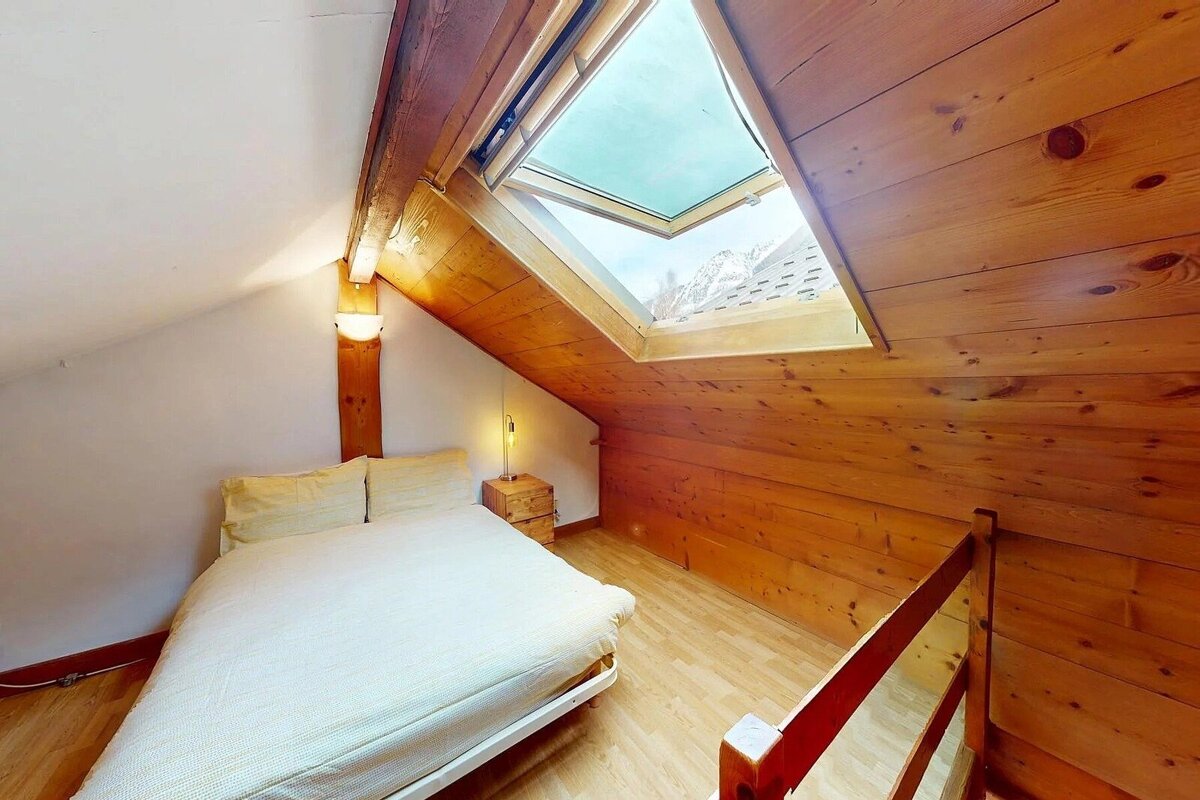 Cozy attic bedroom with warm wooden walls/ceiling, a bright skylight showing a mountain view, and a simple bed with light bedding.
