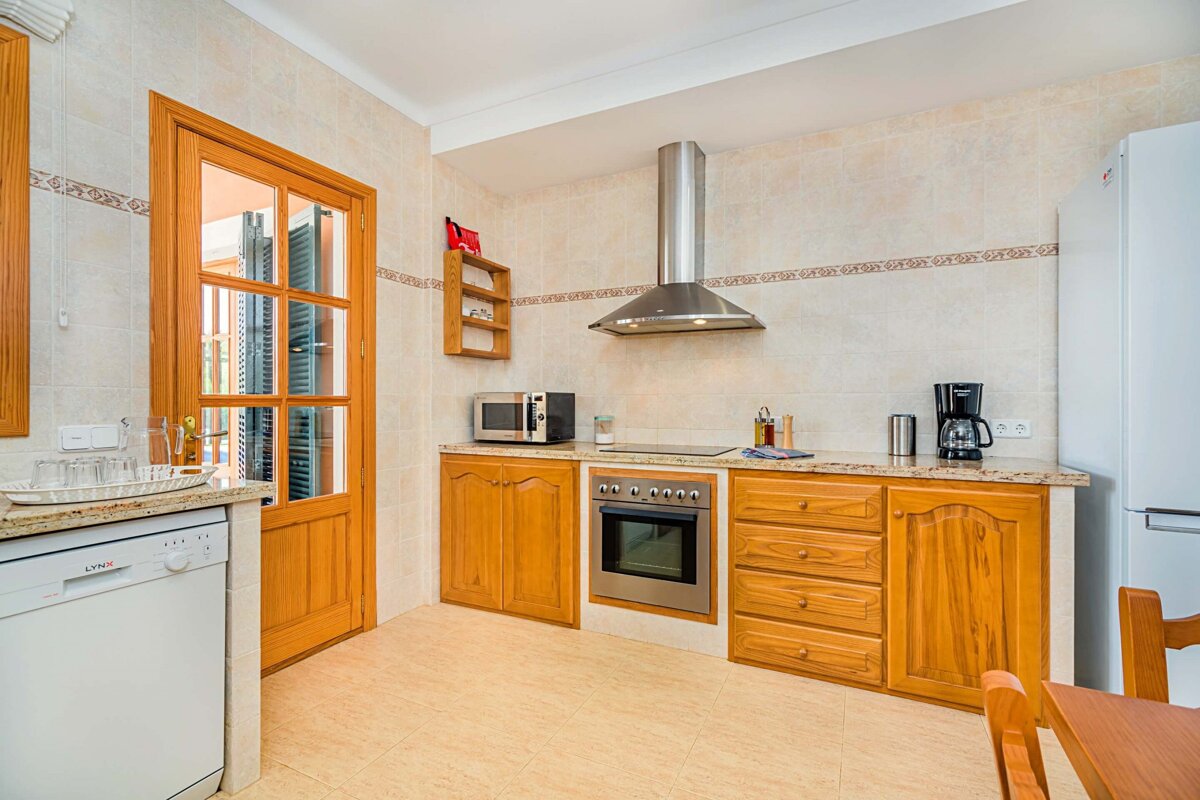 A bright, tiled kitchen with light wood cabinets, modern appliances (oven, microwave, fridge, dishwasher), and a glass-paneled door leading outside.