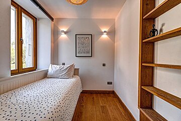 A narrow room with a single bed, polka dot bedding, and wooden floor. White walls feature a window, wall lights, a motivational poster, and wooden shelves.