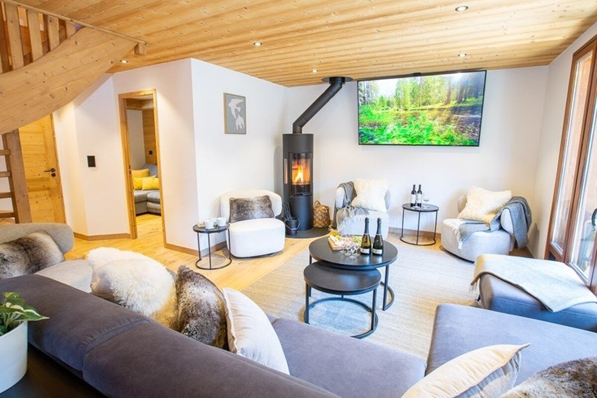 Cozy modern chalet living room featuring wooden ceilings, a wood-burning stove, plush seating, and a large wall-mounted TV.