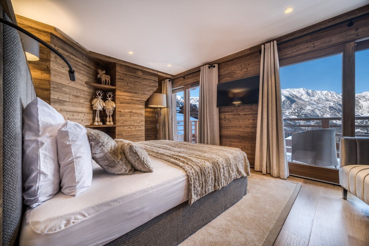 Cozy chalet bedroom with wood walls, a comfy bed, and a breathtaking snowy mountain view from the balcony. Festive deer decor adds charm.