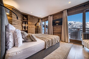 Cozy chalet bedroom with wood walls, a comfy bed, and a breathtaking snowy mountain view from the balcony. Festive deer decor adds charm.