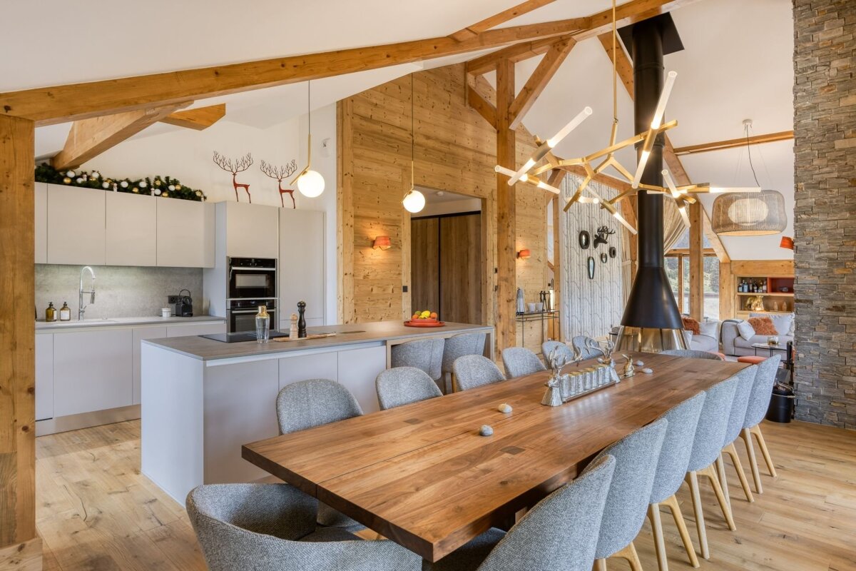 A modern, open-plan kitchen and dining area in a spacious chalet. Features wooden beams, a long dining table, striking chandelier, and subtle holiday decor.