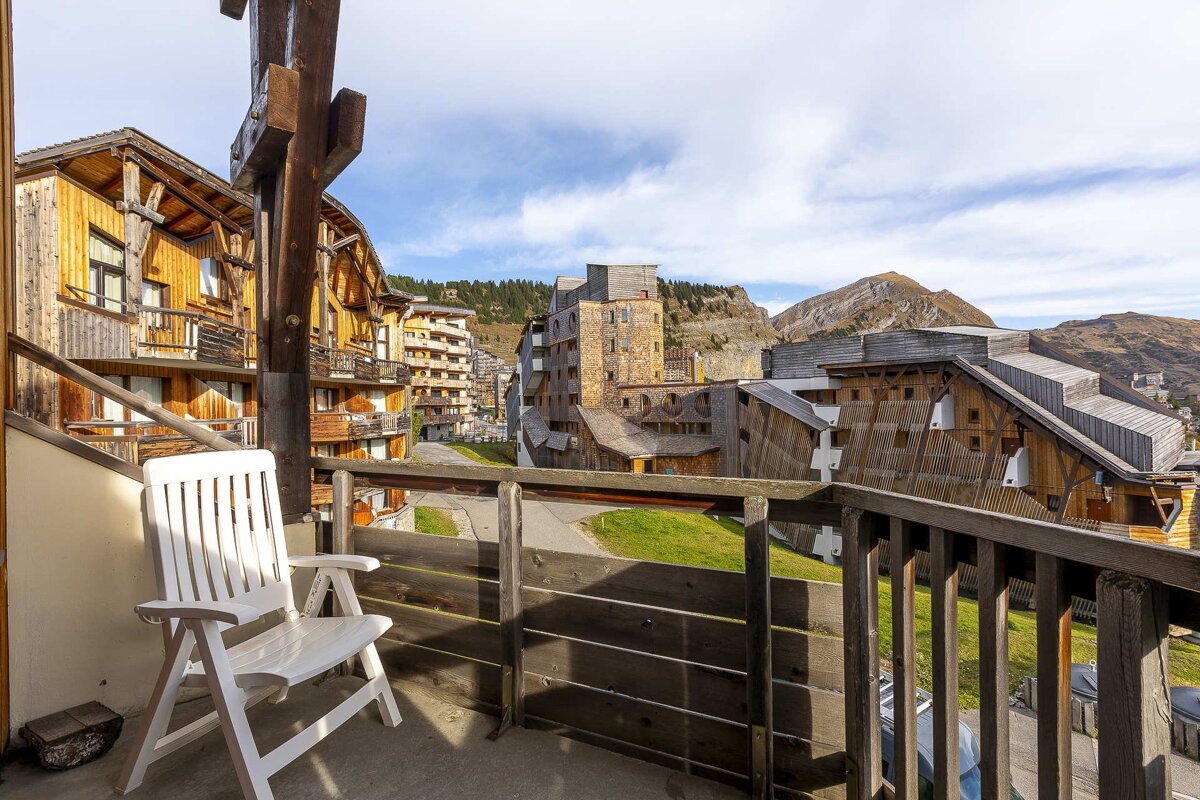 A balcony with a white chair overlooks a mountain village of wooden chalets and distant peaks under a partly cloudy sky.