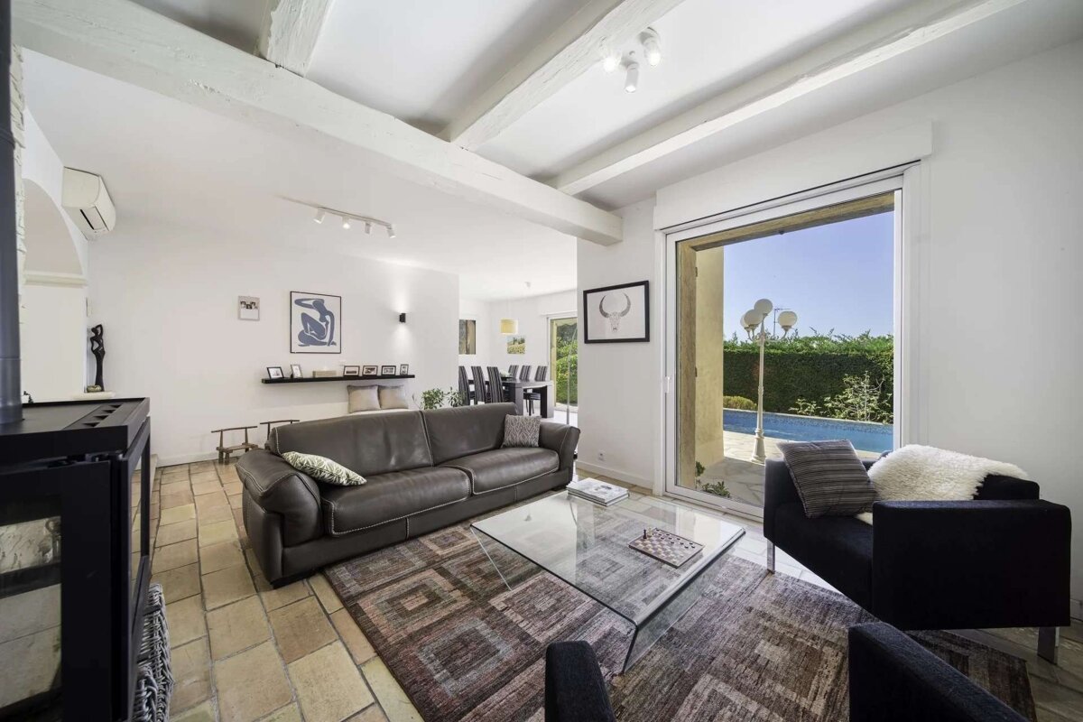 A bright, rustic-modern living room with exposed ceiling beams, dark furniture, and a glass table, leading out to a sunny pool area.