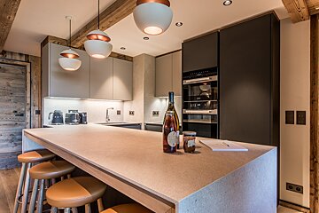 A chic, modern kitchen with a large island, bar stools, sleek appliances, and rustic wooden beams. Unique white and copper pendant lights hang overhead.